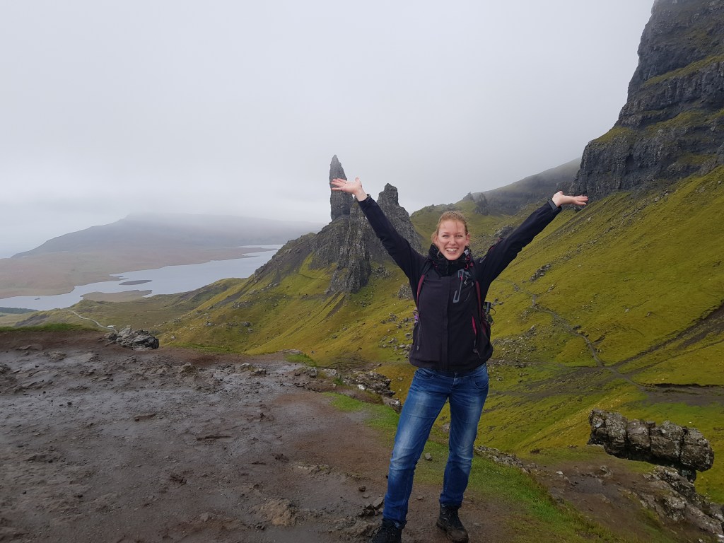 Photo of Christine Kammerer, the author of the article, in the Isle of Skye, Scotland. On the background, the rocky feature called Old Man of Storr is visible. It is a misty and humid day.