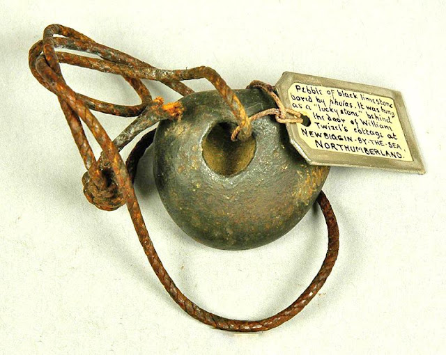 Photo of a stone with a hole and string. A tag attached to the object reads:

"Pebble of black limestone bored by pholas. It was hung as a lucky stone behind the door of William Twizel's cottage at Newbiggin-By-The-Sea, Northumberland.